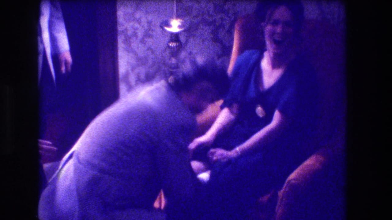 Man in light gray shirt leans toward a laughing woman in a dark blue dress seated in an armchair in a dimly lit room.
