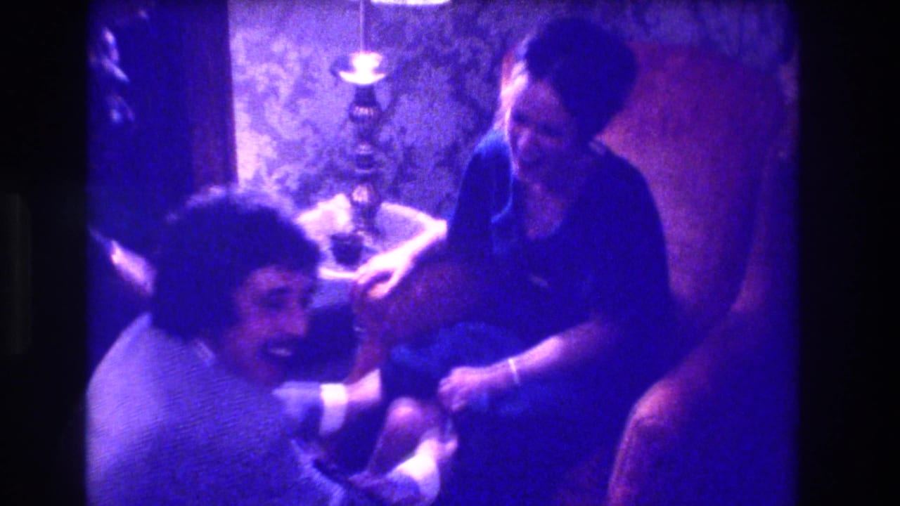 Close view of a dark-haired man leaning over a seated woman in a private home interior, consistent with a 1973 New Jersey domestic gathering; purple 8mm color drift visible.