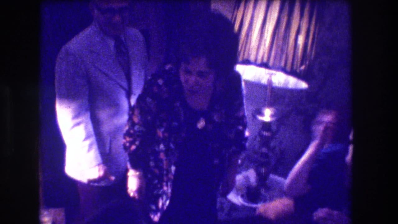 Man in light jacket and woman in dark floral dress stand together in purple-tinted living room beside a lit lamp