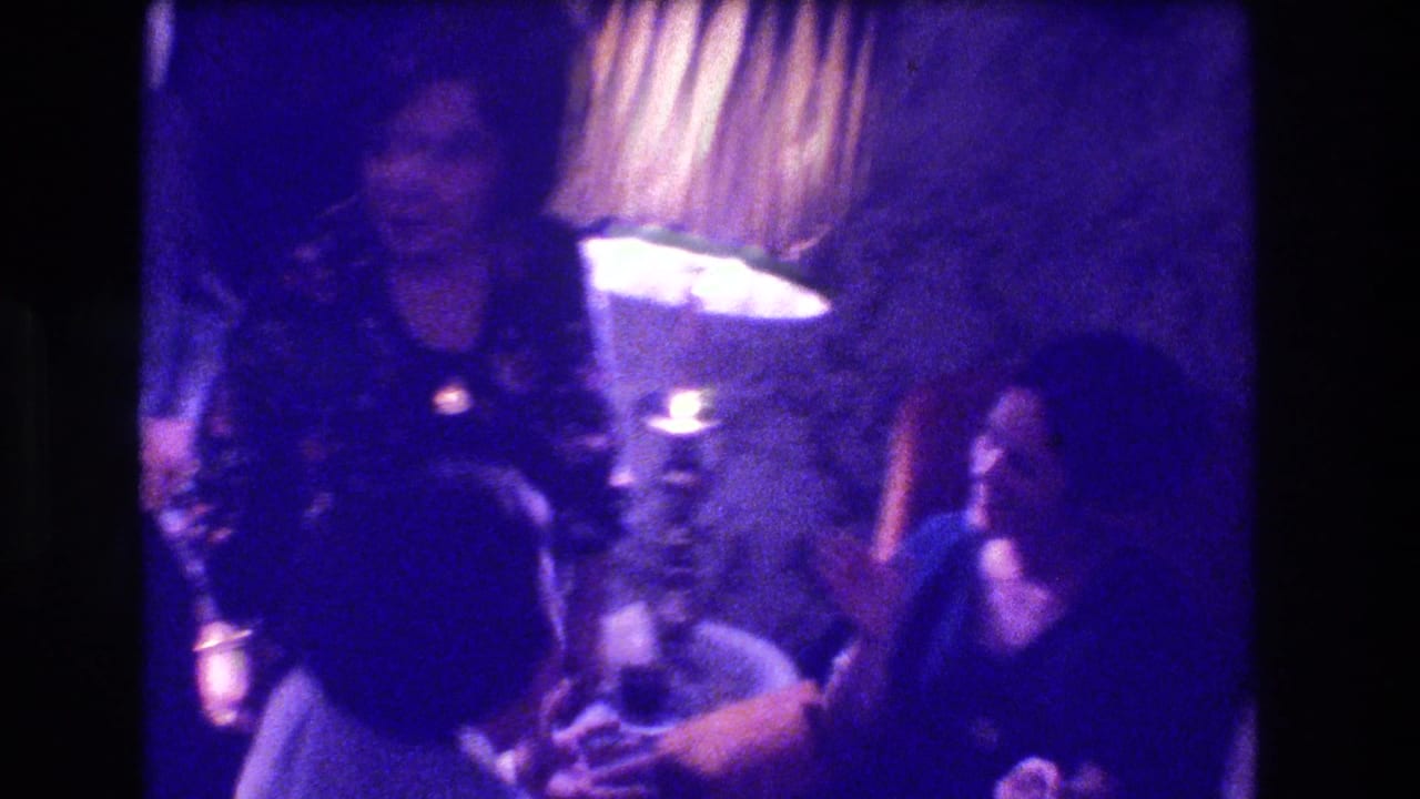 Group of adults in purple-cast living room, woman in dark floral dress at center, lamp visible to right