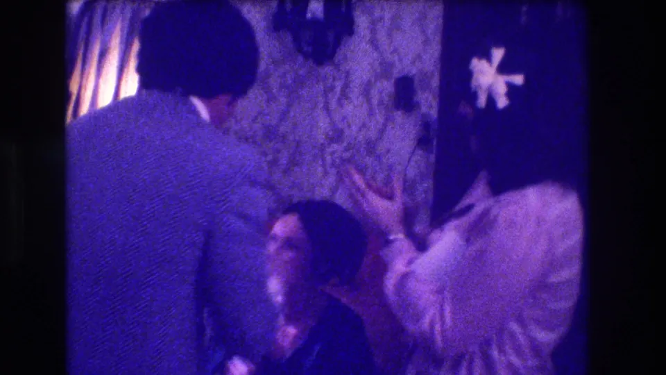8mm home movie, circa 1973, Medford NJ: figure kneeling before woman in domestic interior with patterned wallpaper and pleated lamp shade visible