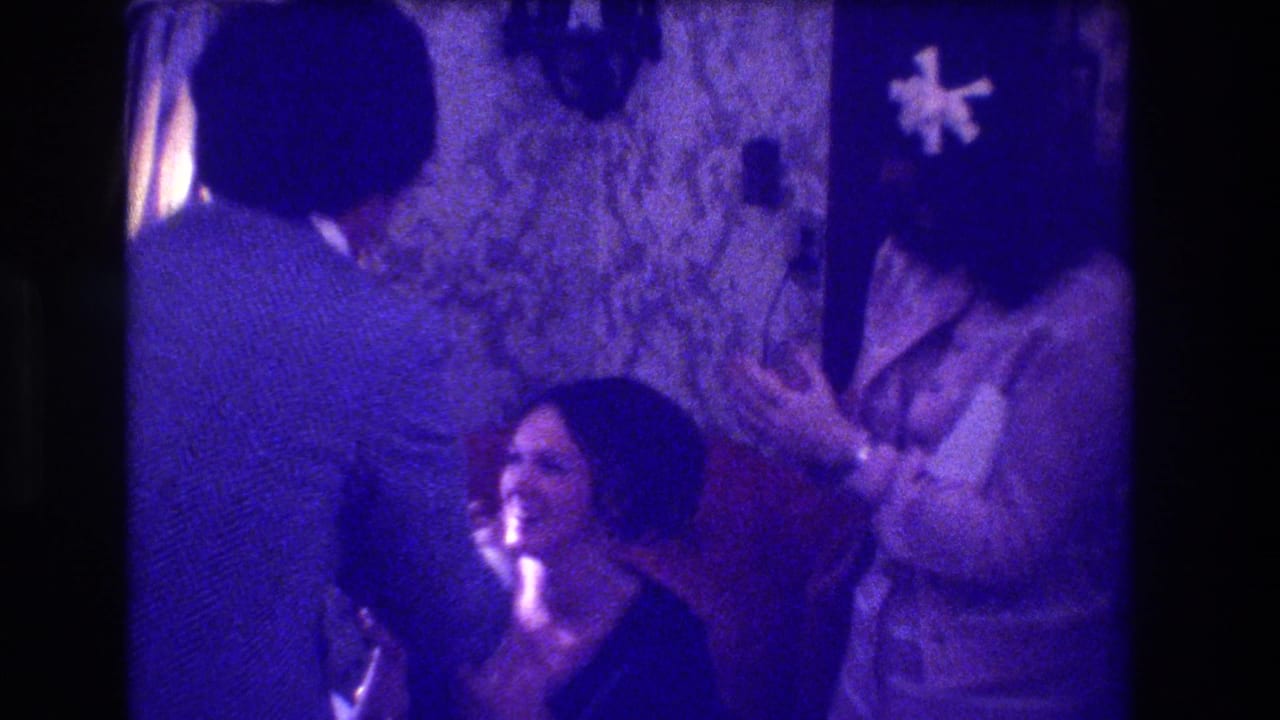 Three adults at an indoor gathering; woman laughing in foreground, another with white flower corsage visible behind her; floral wallpaper; heavy purple 8mm colour cast.