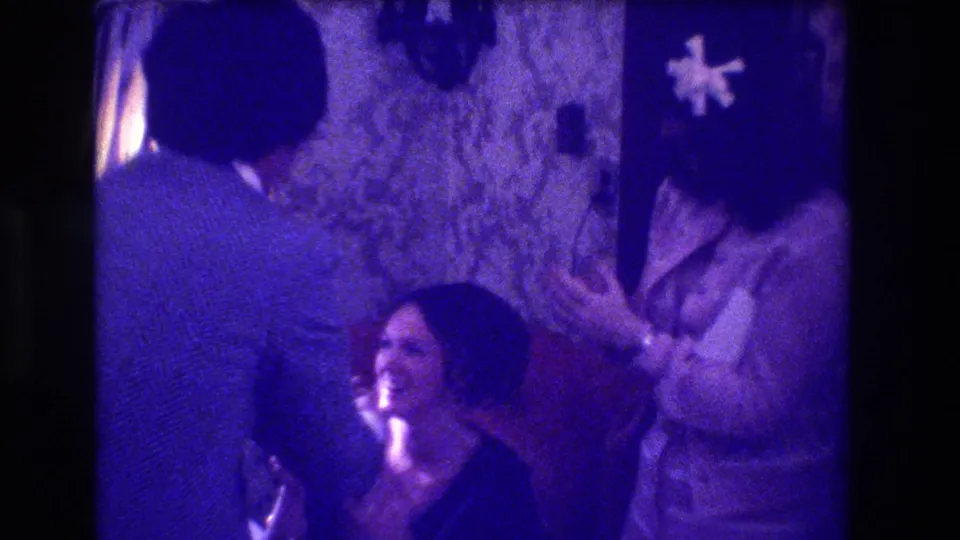Three adults at a 1973 indoor celebration in Medford, New Jersey; laughing woman in foreground, another with white flower corsage; floral wallpaper; heavy purple 8mm colour cast.