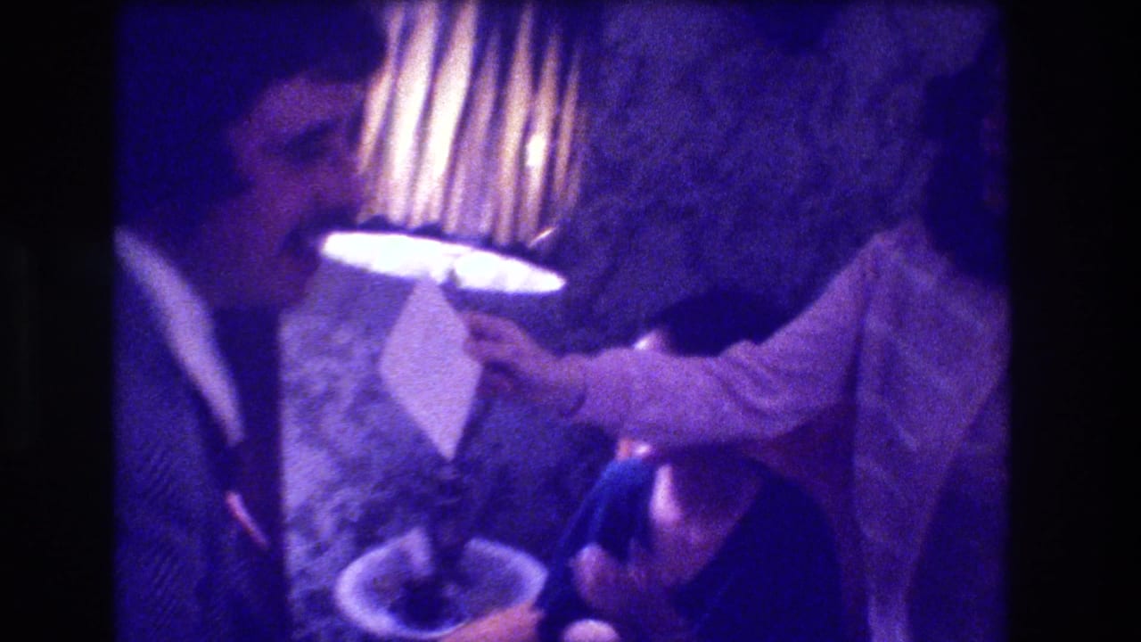 Man leans forward near a pleated lamp, possibly presenting or reading something; 1973 indoor celebration, Medford, New Jersey; plate or bowl in frame; strong purple 8mm colour cast.