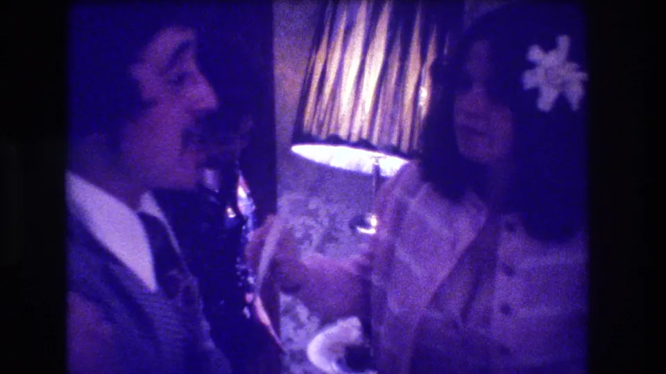 Smiling man in dark suit and woman in satin blouse stand close near a pleated lamp; white flower corsage visible; 1973 indoor celebration, Medford, New Jersey; purple 8mm colour cast.