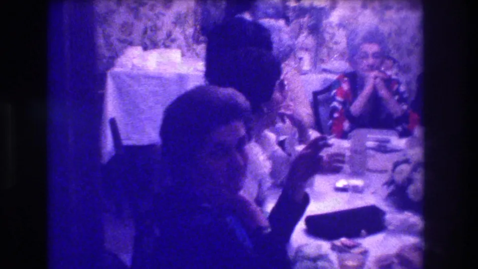 Wider view of a 1973 indoor celebration in Medford, New Jersey, showing a set dining table with dishes, several adults seated and standing, and a woman in a colourful dress at right; purple 8mm colour cast.