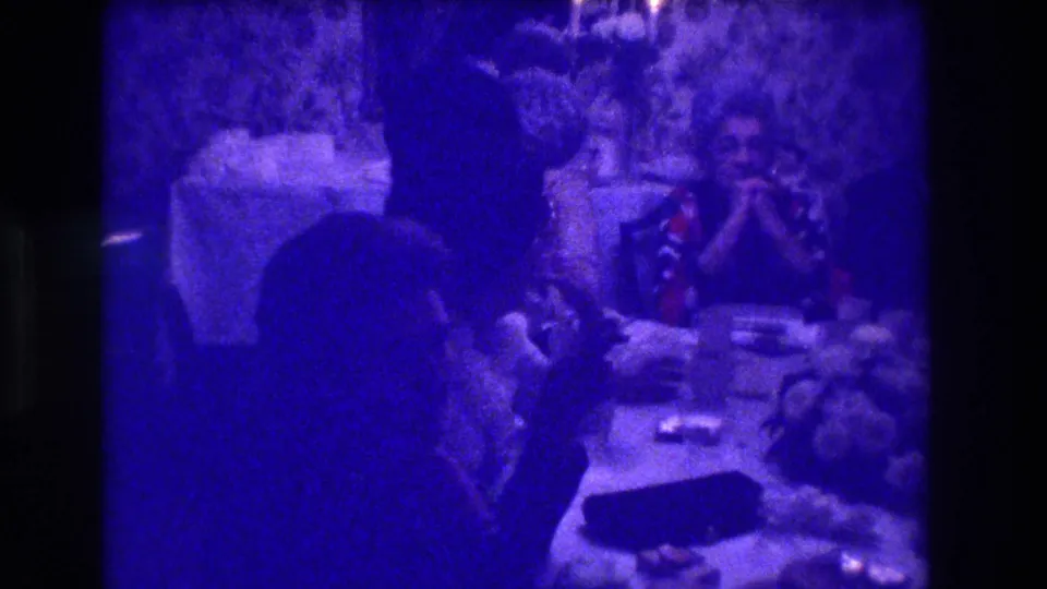 1973 8mm home movie: adults at a dining table in a blue-purple-tinted interior with floral wallpaper, shot past a dark foreground figure, Medford NJ.