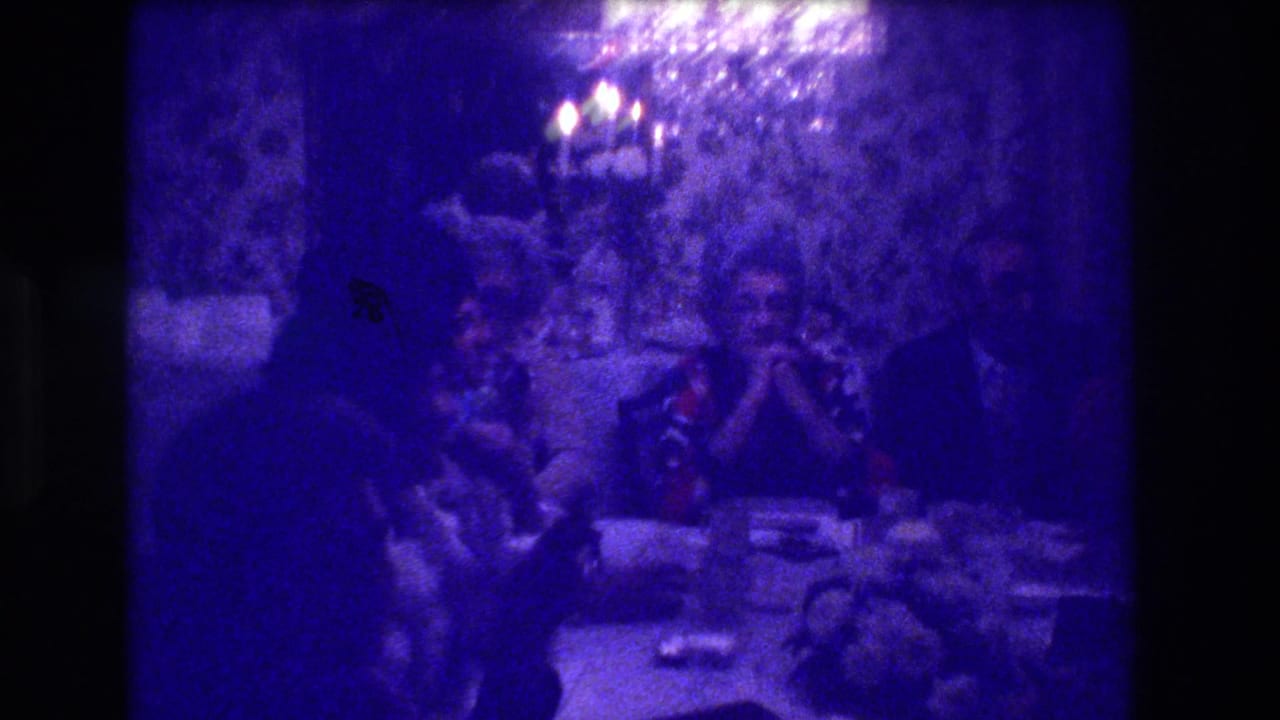 Dining room scene with adults at the table, a dark silhouetted figure in the lower foreground, and a ceiling light source visible in the upper portion.