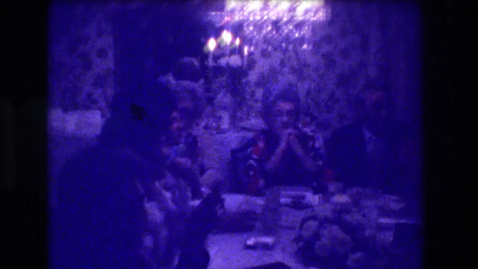 1973 home movie: dining room scene with adults around a set table, heavy blue-purple tint, chandelier light visible at upper right, and a dark silhouette in the foreground.