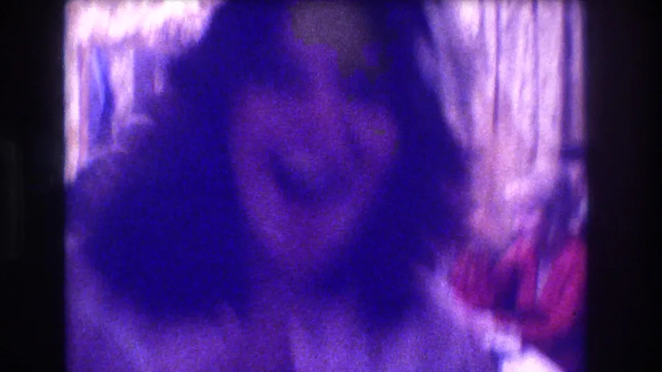 8mm home movie: a person's face and upper body fill the frame in extreme blur and blue-purple tint, with pale curtains to the right; identity indeterminate from this frame.