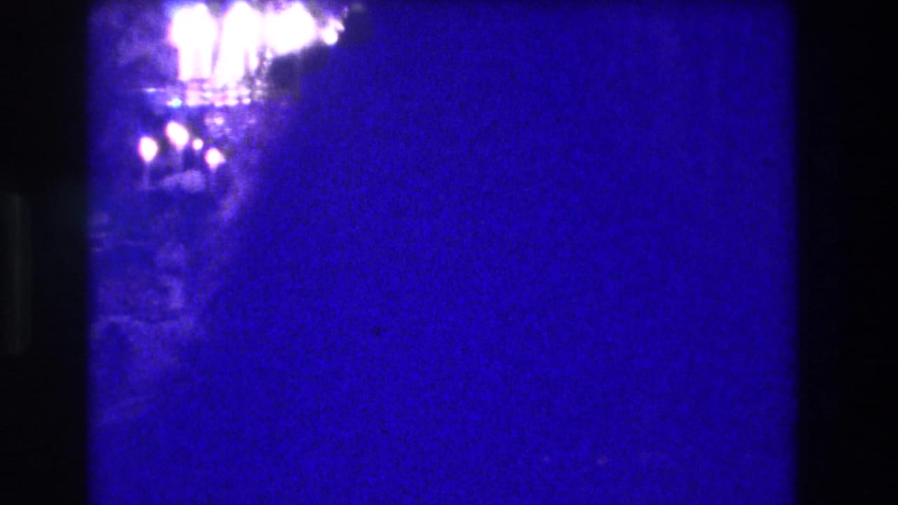 Heavily blue-overexposed 8mm frame with a ceiling light fixture barely visible amid a deep blue-purple color wash.