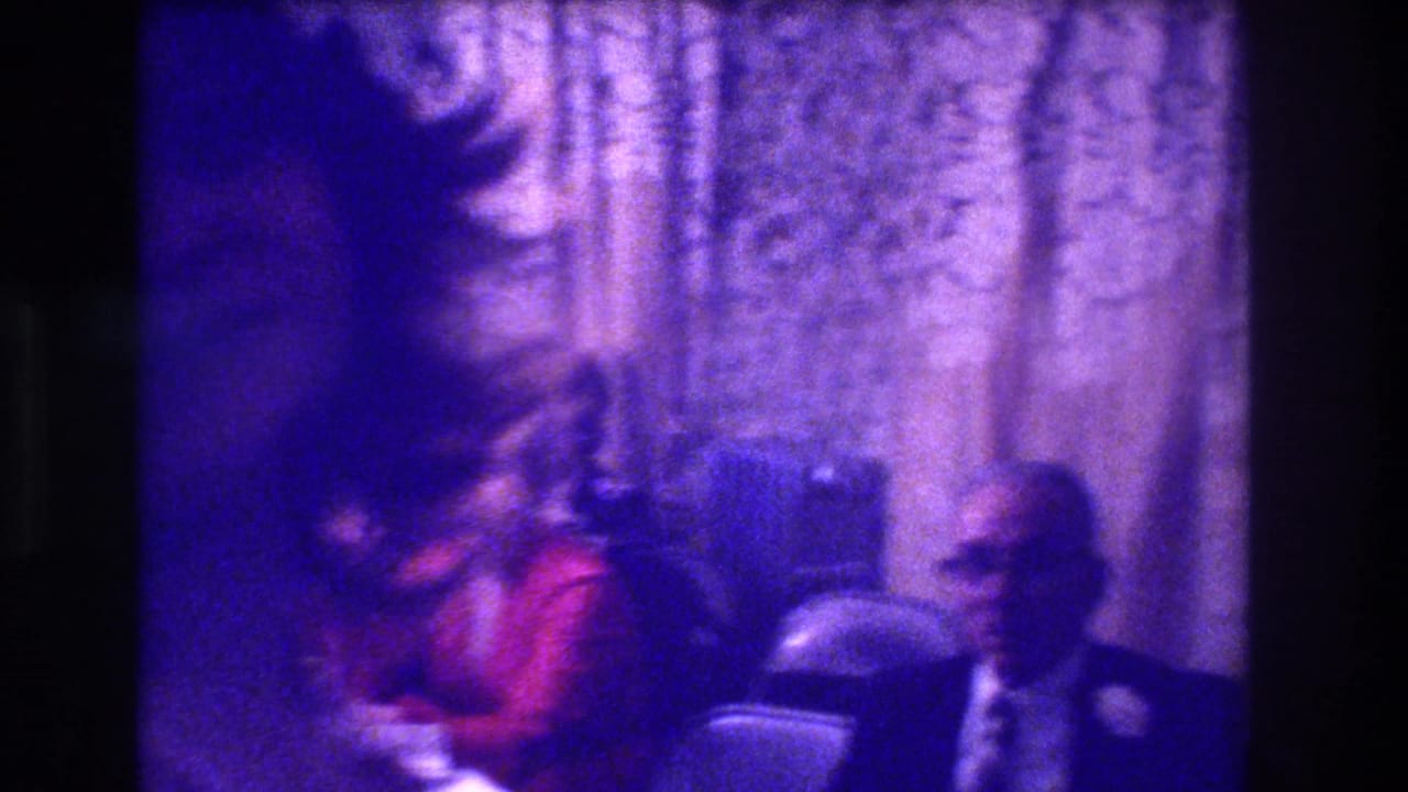 Motion-blurred figure with afro in foreground; woman in red and suited man seated behind gold brocade curtains.