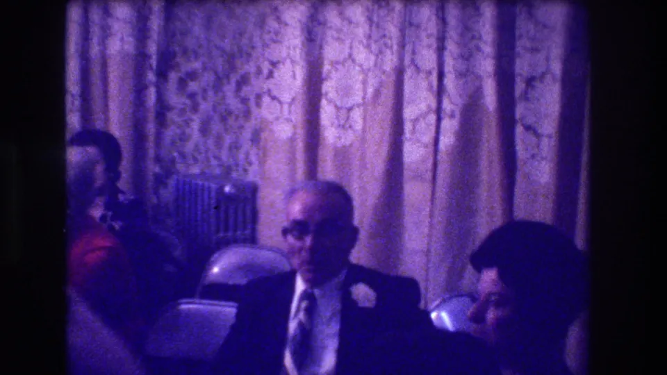 8mm interior frame from a 1973 gathering — a man in a dark suit with white boutonniere seated among adults in folding chairs, gold brocade drapery filling the background.
