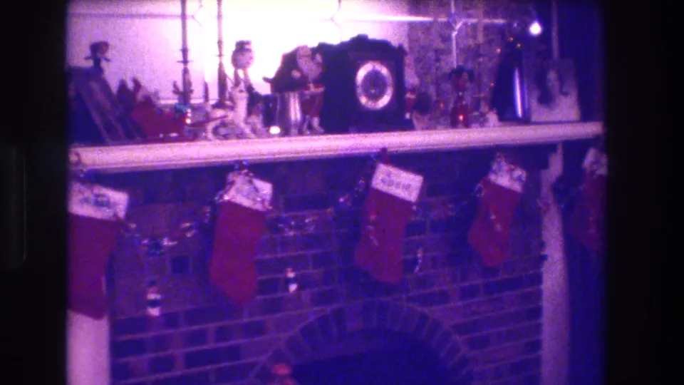 1973 home movie frame of a decorated fireplace mantel with red Christmas stockings bearing name tags, a dark clock, figurines, and tinsel garland in pink-purple color shift.