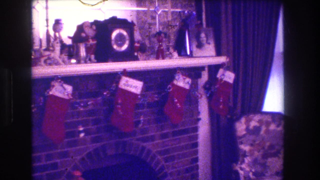 Decorated fireplace mantel with stockings, framed photos, curtains, and a floral chair at the side.