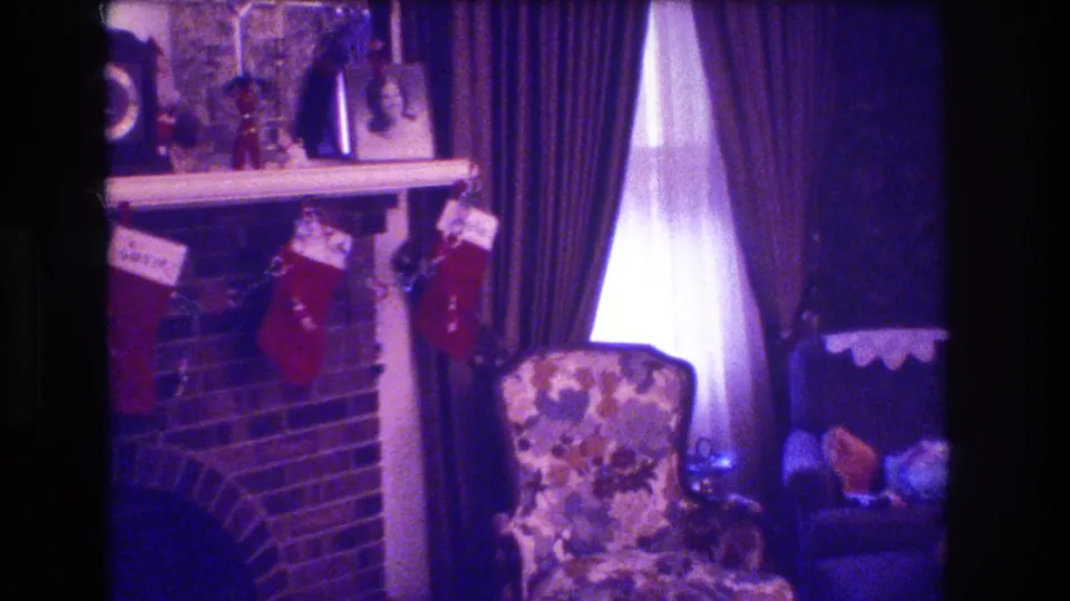 1973 Medford, New Jersey frame showing the right side of a Christmas-decorated fireplace with stockings, heavy curtains, a floral chair, and a small table with household items.