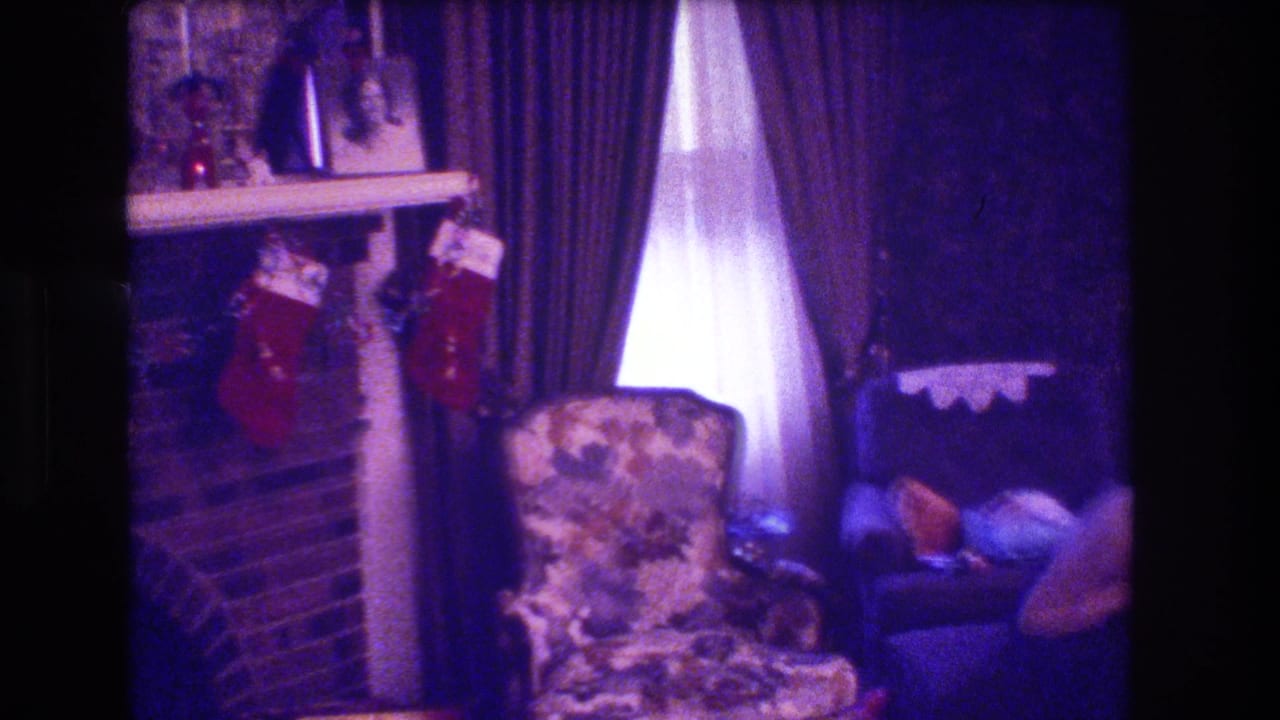 Christmas living room with fireplace stockings, floral chair, curtains, and a woman partly visible at the right edge.