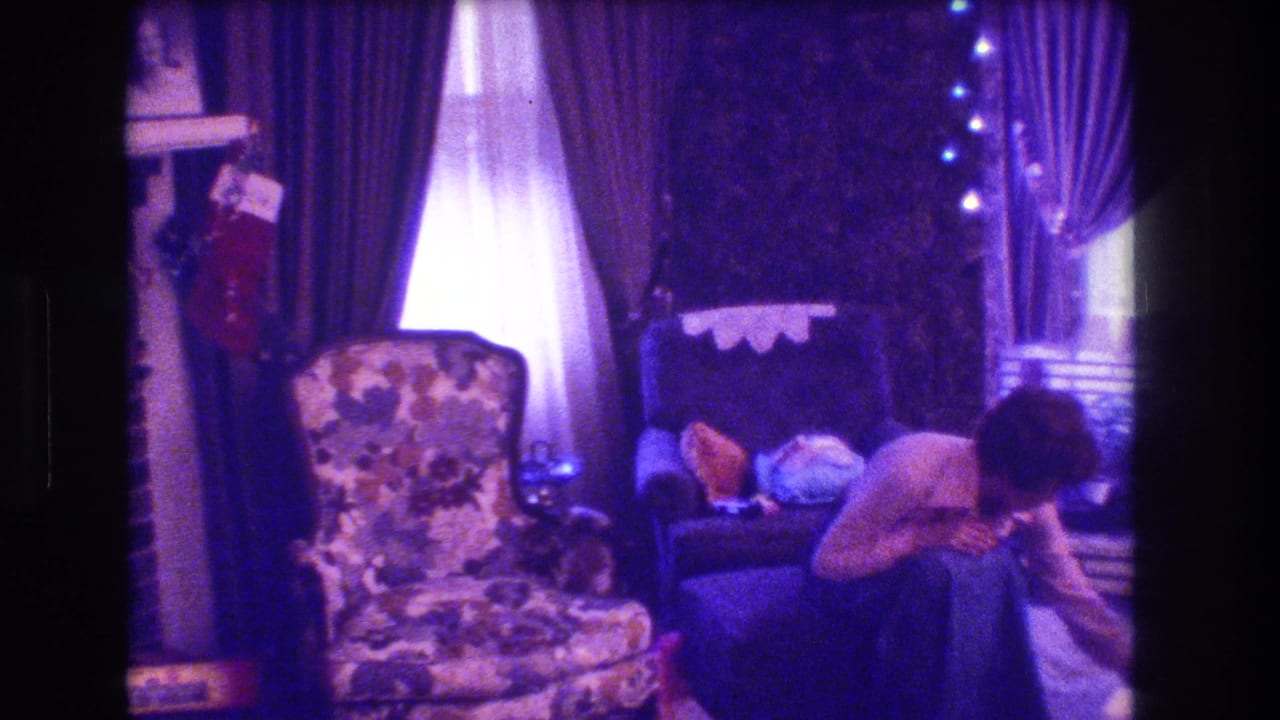 Woman bending near a chair in a Christmas-decorated living room with fireplace, stockings, curtains, and armchairs.