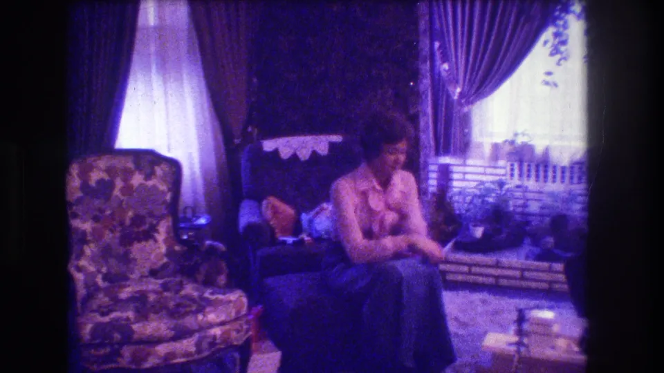 1973 Medford, New Jersey 8mm frame showing a woman seated beside a curtained window in a Christmas-decorated living room with floral upholstery, a dark chair, and fireplace stockings nearby.