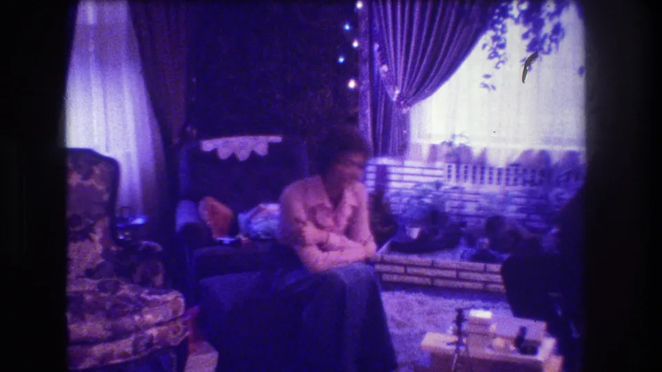 1973 Medford, New Jersey home-movie frame of a woman seated near a bright curtained window, looking toward the camera amid Christmas stockings, upholstered chairs, and a brick fireplace setting.