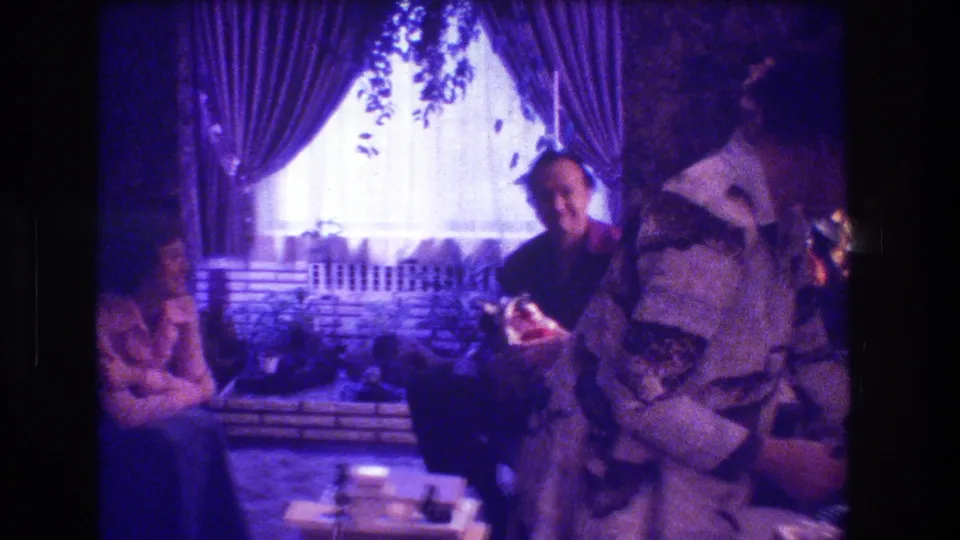 During the Medford, New Jersey Christmas home movie, adults sit and stand around a low table as a person in a light top handles a gift or object near the foreground.