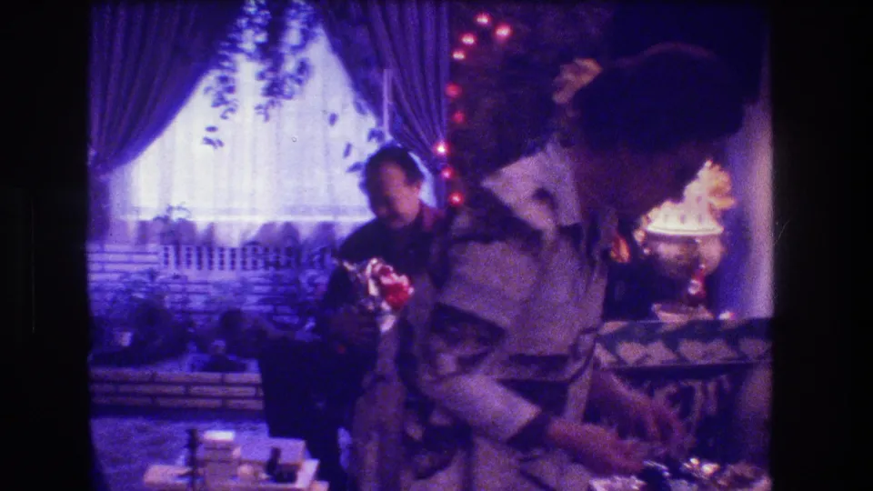 A foreground adult leans over the low table while another adult sits behind them, with the curtained window and decorative lights visible in the 1973 Medford Christmas interior.