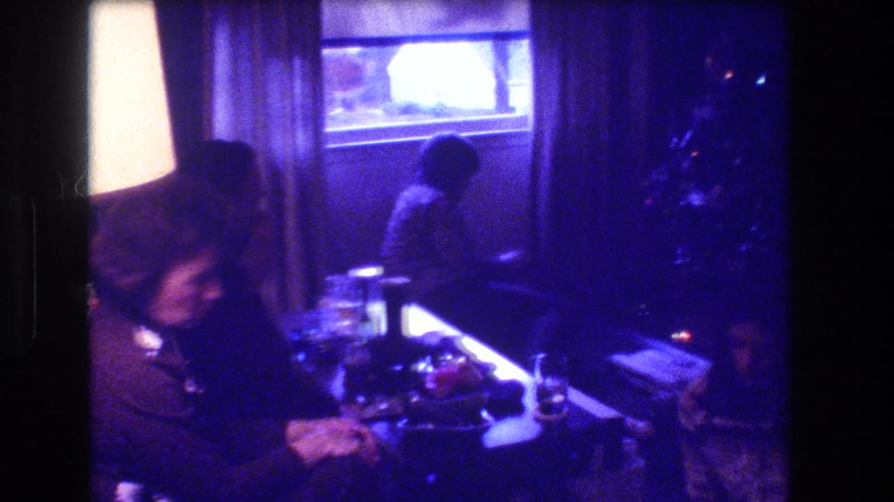 1973 Medford, New Jersey home-movie frame showing adults seated in a dim private living room with a bright table lamp, curtained window, glassware, and holiday decor nearby.