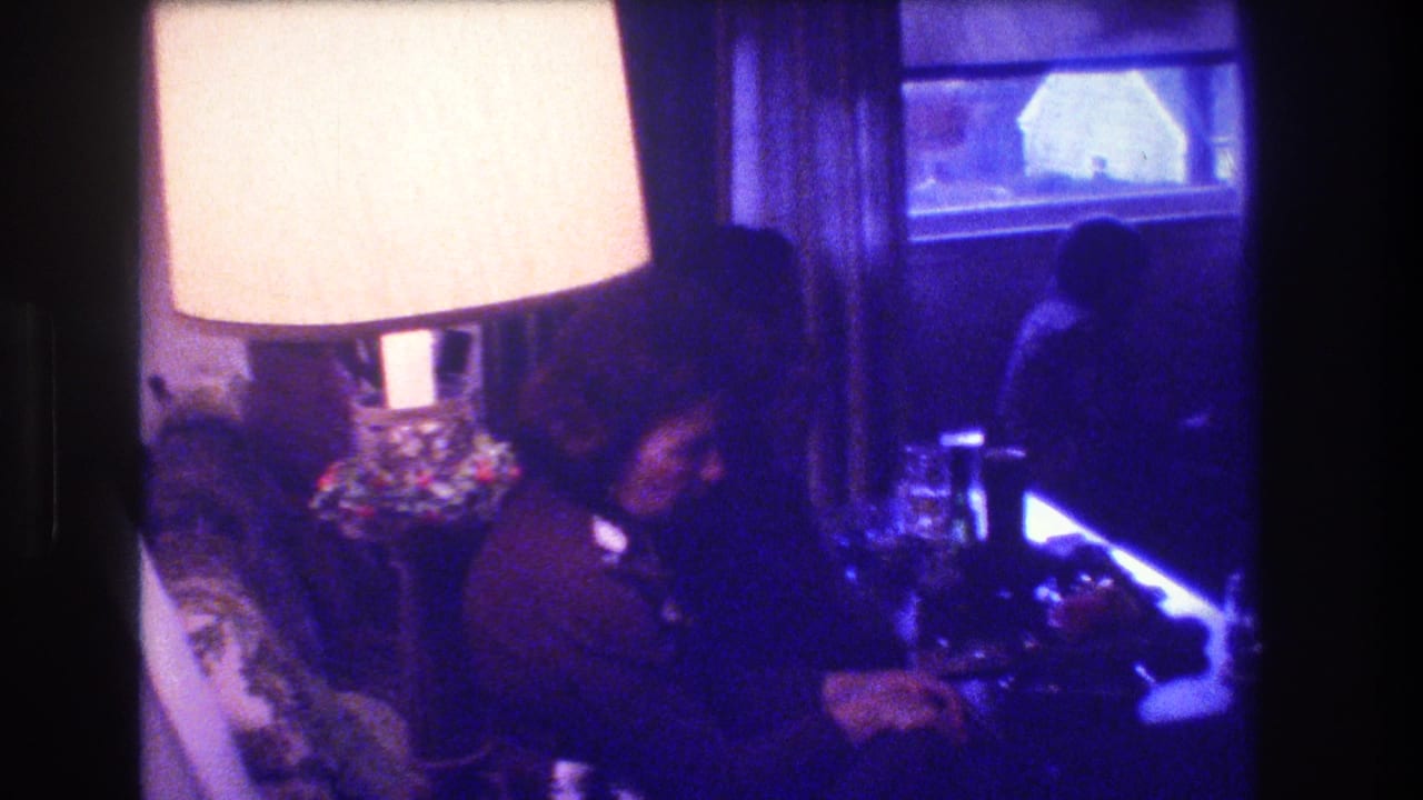1973 Medford, New Jersey home-movie frame showing a seated adult beside a large glowing lamp, with a curtained window and dim living room furniture in view.