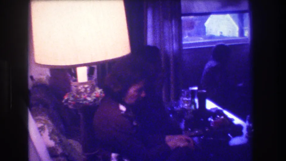 1973 Medford, New Jersey home-movie frame showing a seated adult beside a large glowing lamp, with a curtained window and dim living room furniture in view.