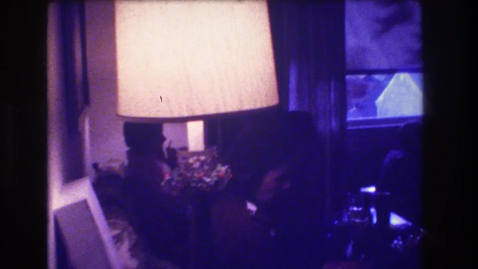 1973 Medford, New Jersey home-movie frame centered on a large lit table lamp, with adults partly visible in a dim living room near a curtained window and holiday decoration.