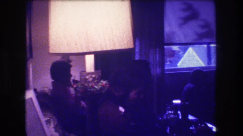 1973 Medford, New Jersey home-movie frame showing an adult in profile seated near a bright lamp, curtained window, and small Christmas tree inside a private living room.