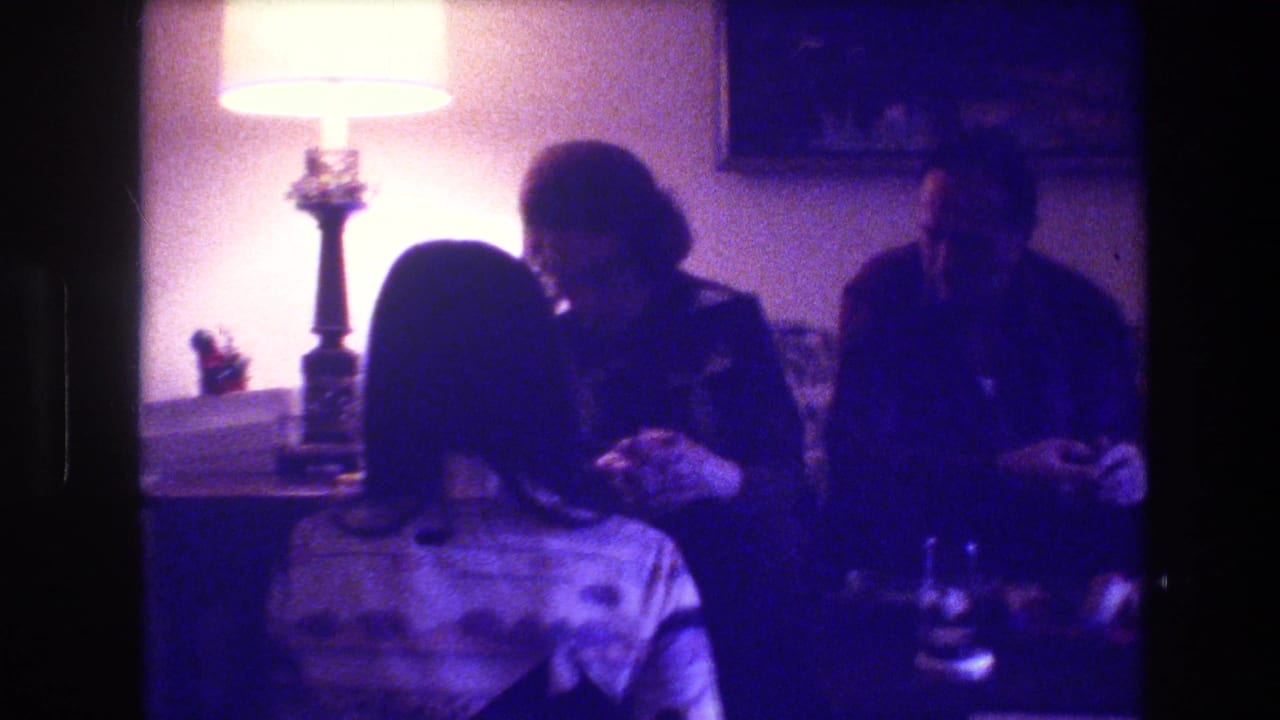 In 1973 Medford, New Jersey home-movie footage, adults sit closely on a couch in a dim living room with a bright table lamp and framed wall art behind them.
