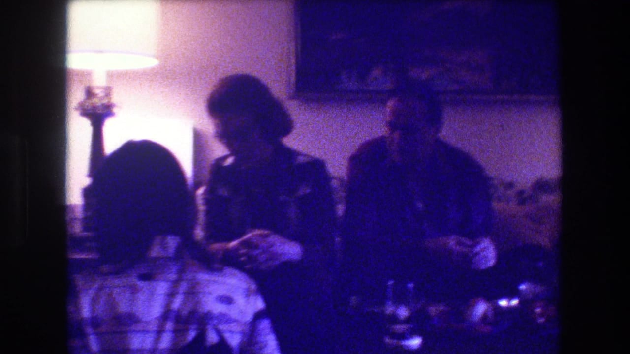 Three adults appear seated together on a couch in the Medford, New Jersey living room, with soft 8mm color shift, a lit lamp, and table items visible in front.