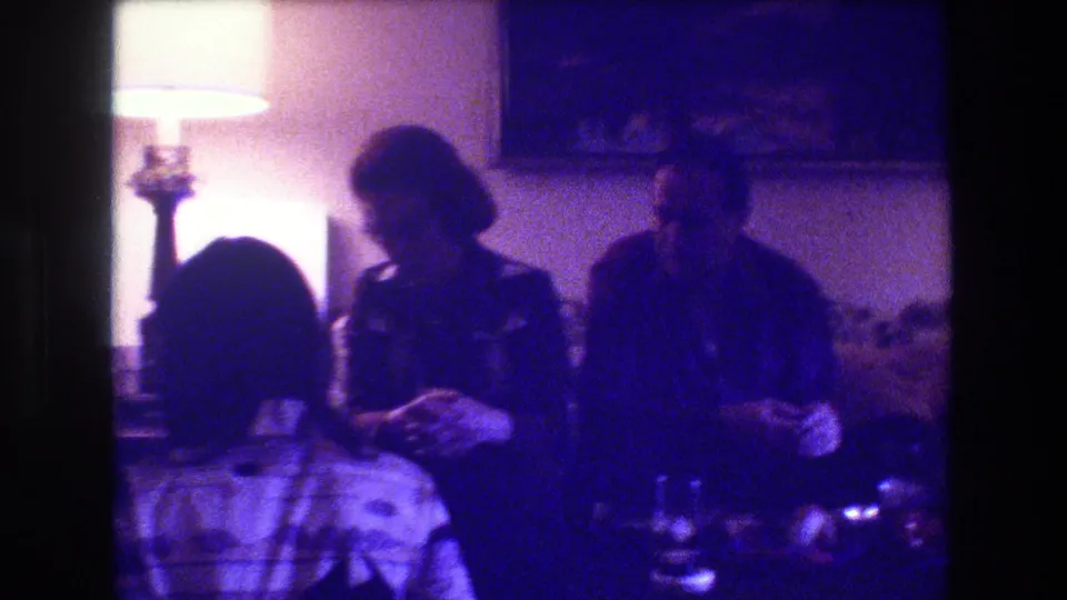 Three adults appear seated together on a couch in the Medford, New Jersey living room, with soft 8mm color shift, a lit lamp, and table items visible in front.