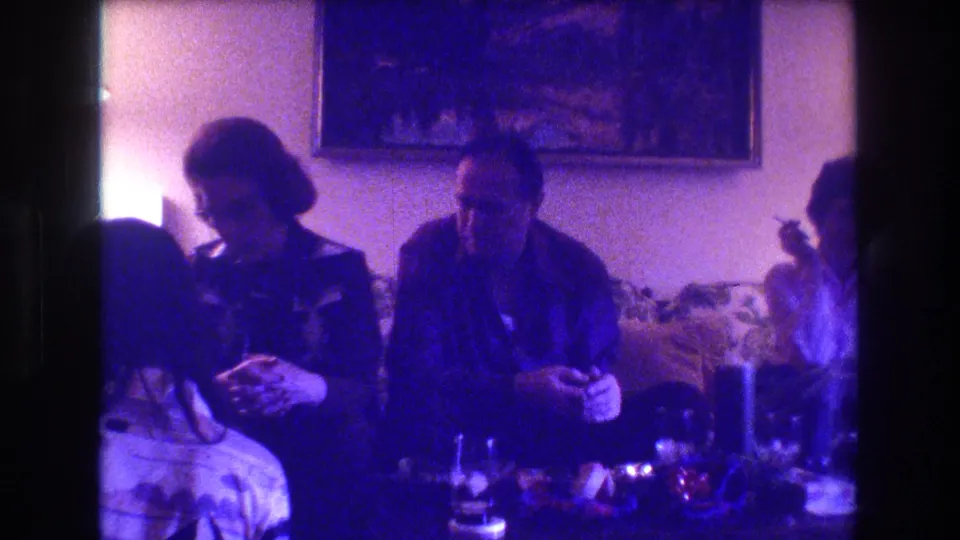 The home-movie frame shows adults gathered on a couch in a private Medford living room, with one person partly turned away and drinks or table items in the foreground.