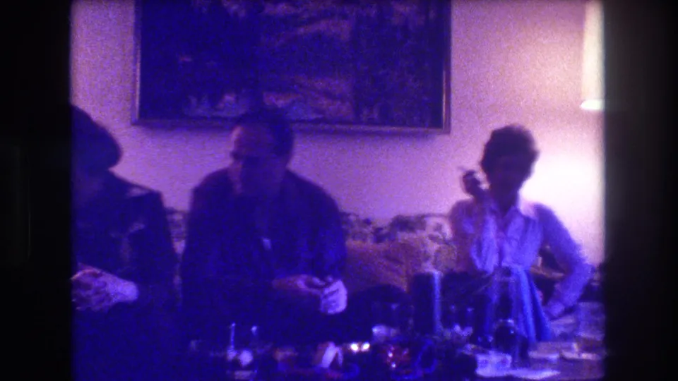In the final frame, adults remain seated in the dim 1973 Medford living room as a person at right appears to hold a cigarette beside the lamp and tableware.