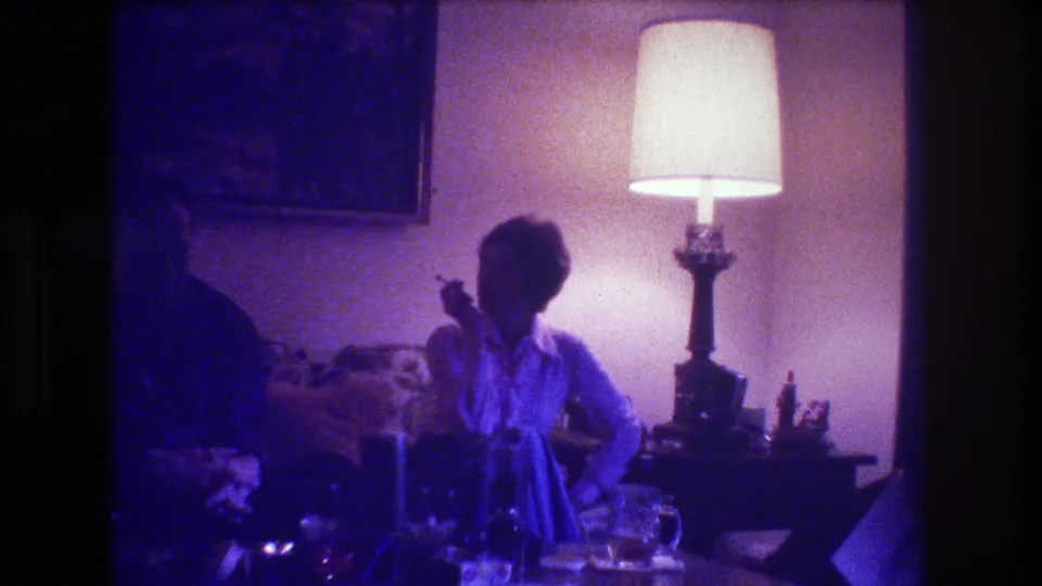 Medford, New Jersey 1973 home-movie frame showing the woman smoking near the center, with the man mostly obscured at left, a lamp behind her, and drinks on the table.