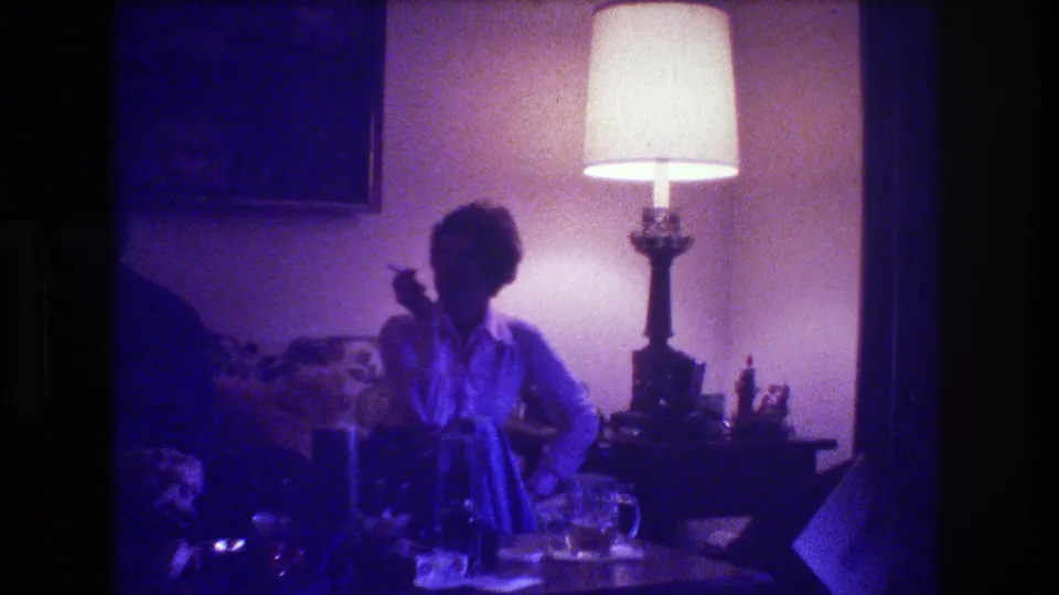 Medford, New Jersey 1973 frame showing a woman seated on a couch with a cigarette raised, a bright table lamp behind her, and a darkened living-room table in front.