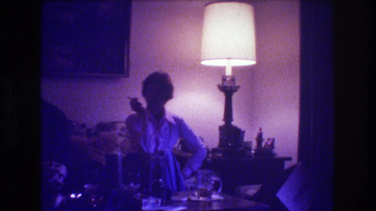 Woman seated on a couch beside a lit table lamp, holding a cigarette in a dim living room.
