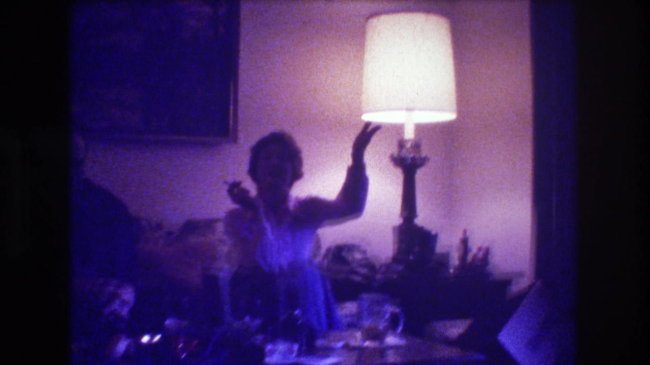 Woman on couch raises one hand near a lit lamp while holding a cigarette.
