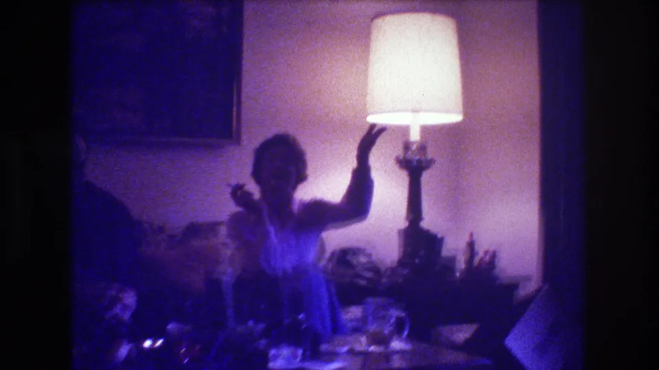 The seated woman appears to wave or gesture toward the camera beside a bright table lamp in the purple-cast 1973 Medford living-room footage.