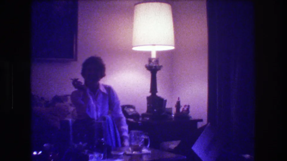 A woman sits in partial profile on a couch, cigarette in hand, with table glassware in the foreground and a glowing lamp defining the private 1973 living-room interior.