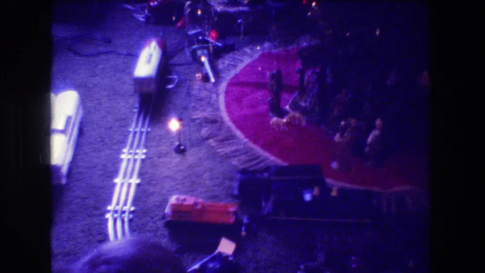 1973 Medford, New Jersey holiday home-movie frame with train tracks, a red circular rug, tree ornaments, a small lamp, and dark train cars near the lower edge.