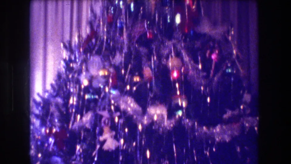 1973 Medford, New Jersey frame of a holiday tree indoors, with silver tinsel, ornaments, colored lights, and pale curtains in the background.