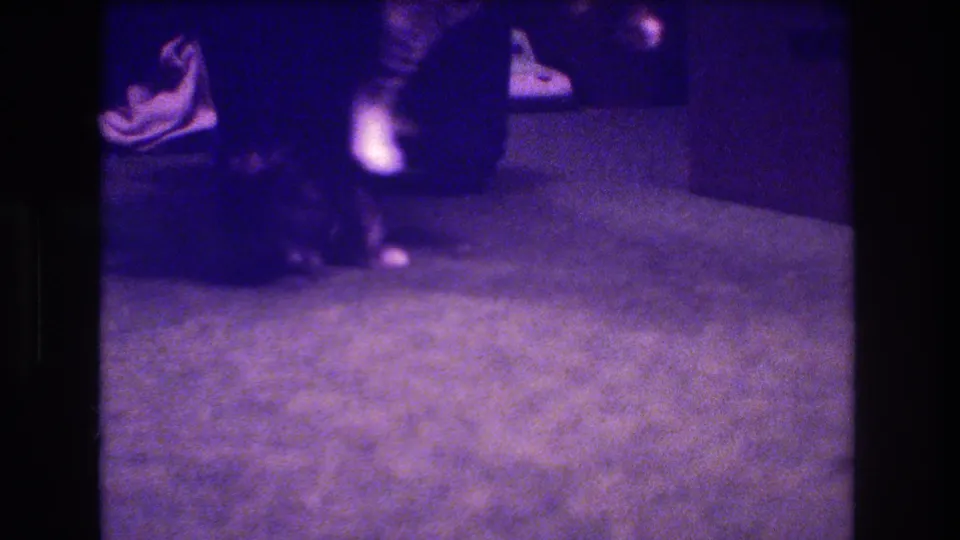 A dark kitten stays low on the carpet near an adult foot in a dim, grainy 8mm home-movie frame associated with Medford, New Jersey, 1973.