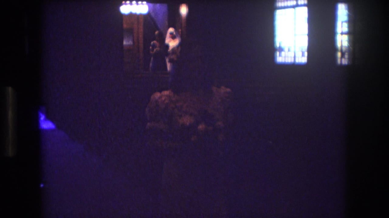 1974 Boston 8mm home-movie frame showing a dim church wedding scene with a veiled bride near the altar, glowing windows, and guests mostly in shadow.