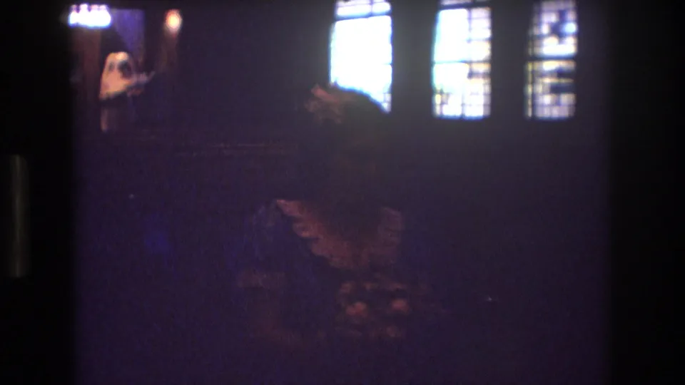 1974 Boston home-movie frame showing a dark church wedding ceremony, with the veiled bride near the altar and bright side windows cutting through the underexposed scene.