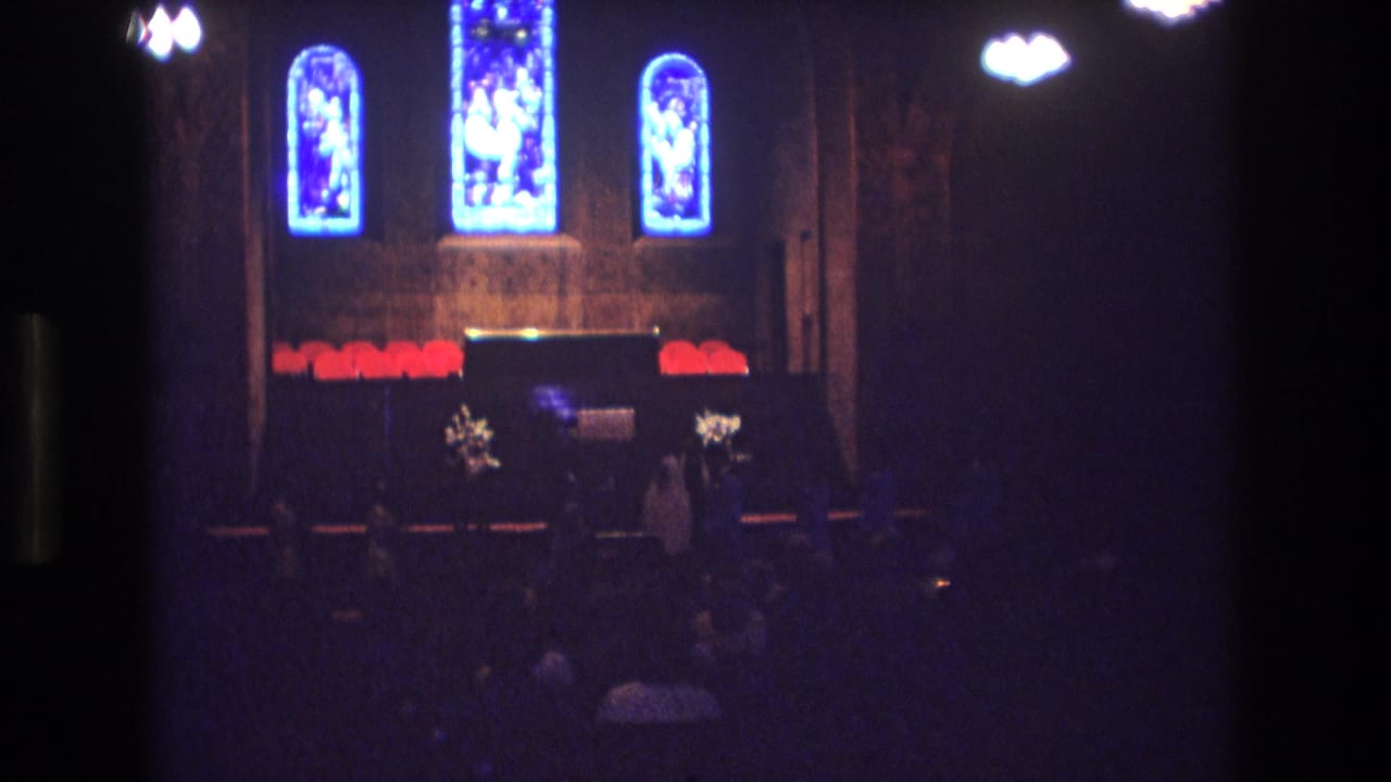 1974 Boston 8mm frame of a church altar area with glowing blue stained glass windows, red altar decorations, floral arrangements, and dark pews in low interior light.