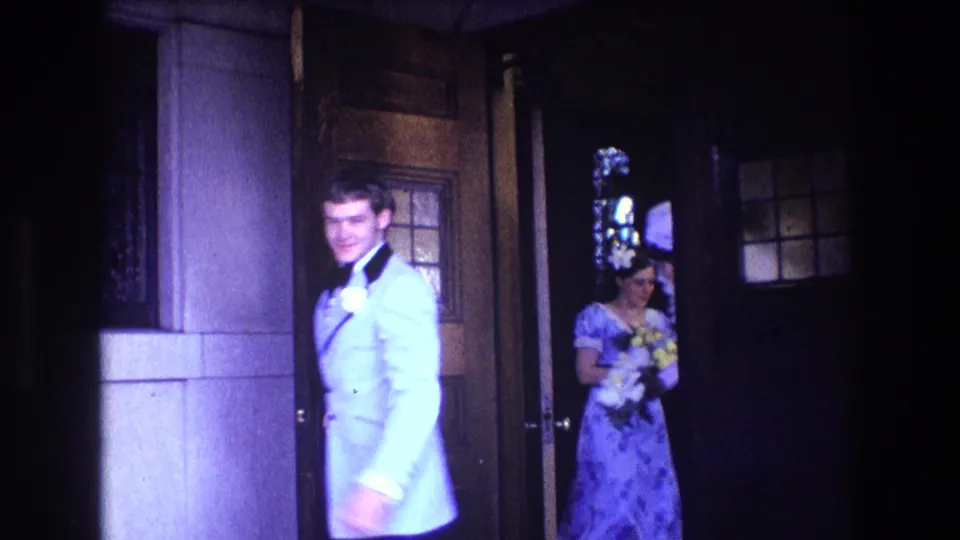 1974 Boston 8mm frame showing a groom in a light-colored tuxedo leaving the church doorway, with a bridesmaid or attendant in blue visible behind him carrying flowers.