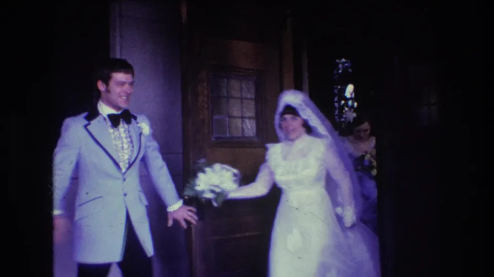 1974 Boston 8mm frame of the bride in a white gown and veil exiting the church beside the groom in a light tuxedo, with attendants and flowers visible near the doorway.